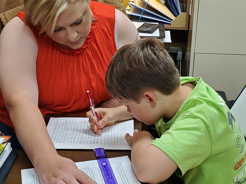A woman looks on while a young boy writes on a piece of paper.