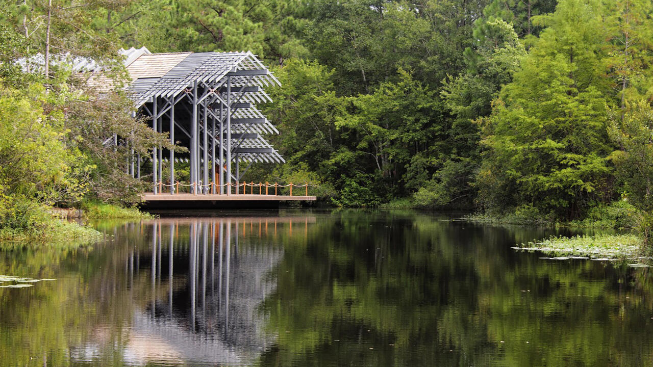 The Pinecote Pavilion, a symmetrical, wooden shed with exposed beams and a broad gable roof, stands across the pond at the Mississippi State University Crosby Arboretum. 